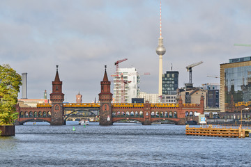 Die Berliner Oberbaumbrücke überspannt die Spree im Morgenlicht mit dem Rathaus, dem Fernsehturm und mehreren Baukränen im Hintergrund - obrazy, fototapety, plakaty
