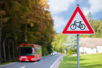 Attention, a red and white triangular road sign warns of cycling that can cross the road. Rural road at the forest with a red bus in the background.
