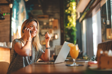 Beautiful young woman talking on the phone in cafe with a laptop on the table in front of her and looking out the window