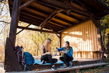 Obraz na płótnie Canvas Two girls in sportswear sitting in woods under canopy and drink tea from thermos