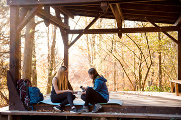 Fototapeta na wymiar Two girls in sportswear sitting in woods under canopy and drink tea from thermos