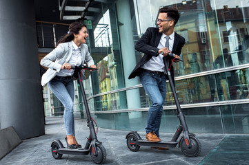 Fototapeta na wymiar Two smiling business people driving electric scooter in front of modern business building going on work.