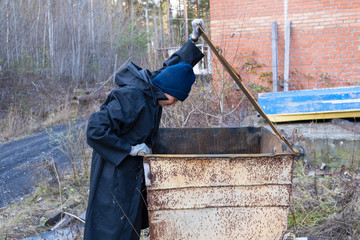 homeless woman in black coat and blue hat is open street garbage cans. woman is rummaging for some food in garbage. poverty.