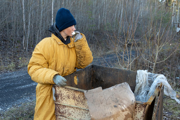 homeless woman in a yellow old ragged jacket and blue hat is eating marshmallows near the open a garbage can