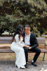 Naklejka premium Couple of love embracing and kissing on bench. Loving couple pre-wedding outdoor photo shoot. Loving romantic couple. Happy girl in white dress and guy posing in suit. Beginning of a love story.
