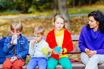 Mother with three children sitting on the bench with some drinks in autumn park.