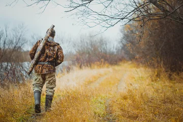 Schilderijen op glas Male hunter walking on the road in the forest in autumn. The gun in the case on the shoulder of the hunter. © nadezhda