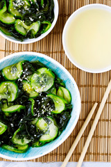 Traditional Japanese dish tasty appetizing seaweed salad served with cucumber in bowl close-up on light blue background top view.Organic natural product.Raw,vegan,vegetarian food.Vertical orientation
