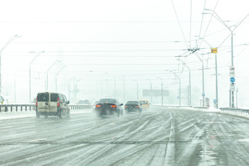Cars drive along road with slush, snowstorm. City traffic in snow blizzard.