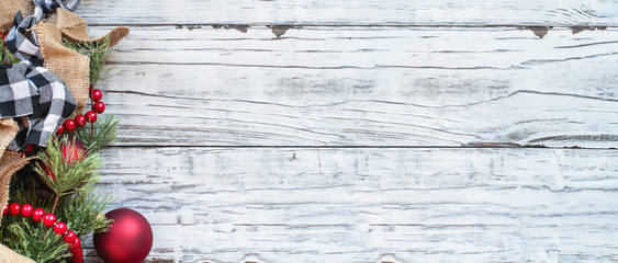 Christmas background banner with holiday trimmings of pine tree branches, ornaments, black and white buffalo check ribbon, burlap and red bead garland. Top view with copy space available.