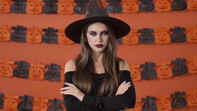 Serious pretty young witch woman in black halloween costume showing silence gesture with finger and crossing her arms over orange pumpkin wall