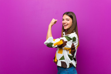 young pretty woman feeling happy, satisfied and powerful, flexing fit and muscular biceps, looking strong after the gym against purple background
