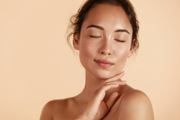 Beauty face. Woman with natural makeup and healthy skin portrait. Beautiful asian girl model touching fresh glowing hydrated facial skin on beige background closeup. Skin care concept - obrazy, fototapety, plakaty