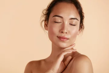 Poster Beauty face. Woman with natural makeup and healthy skin portrait. Beautiful asian girl model touching fresh glowing hydrated facial skin on beige background closeup. Skin care concept © puhhha