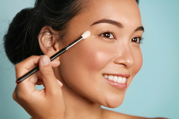 Beauty. Woman applying makeup on eyes with cosmetic brush closeup. Portrait of happy smiling asian...