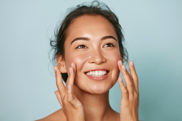 Beauty face. Smiling asian woman touching healthy skin portrait. Beautiful happy girl model with fresh glowing hydrated facial skin and natural makeup on blue background at studio. Skin care concept - obrazy, fototapety, plakaty