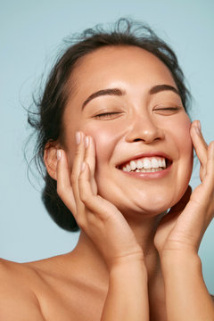 Skin Care. Woman With Beauty Face Touching Healthy Facial Skin Portrait. Beautiful Smiling Asian Girl Model With Natural Makeup Touching Glowing Hydrated Skin On Blue Background Closeup