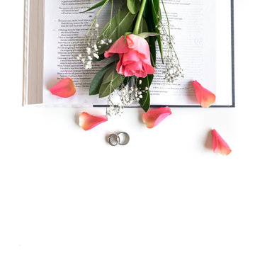 Wedding flat lay: Pink rose, pedals, wedding rings and a Bible on white background 