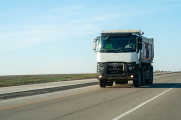 Fototapeta na wymiar A white dump truck drives out of town on a flat asphalt road among fields against a blue sky on a sunny day. A machine for transporting building materials rides on the highway