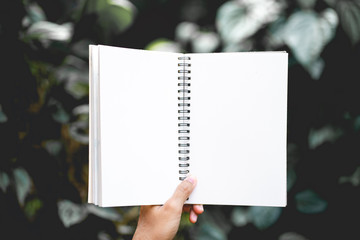 Hand girl holding notebook with blank space for add text on fresh green leaves background with natural texture of seamless foliage. Office workplace.