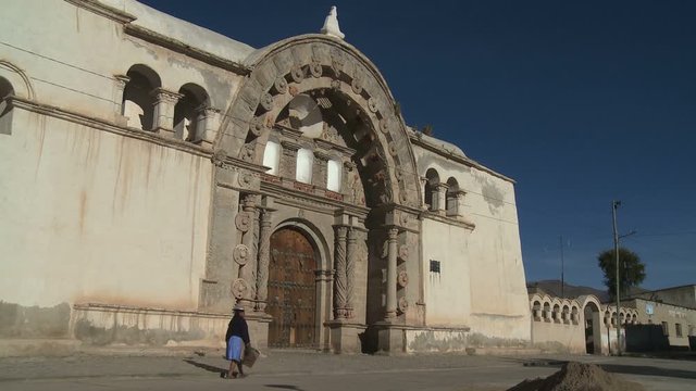 Native Indigenous woman passing by in front of a catholic spanish style church facade in a village in South America