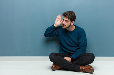 young handsome man sitting on floor looking serious and curious, listening, trying to hear a secret...