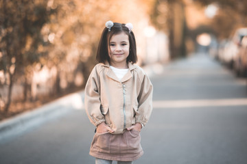 Smiling kid girl 3-4 year old wearing stylish jacket and skirt posing over city background outdoos. Looking at camera. Childhood. Autumn season.