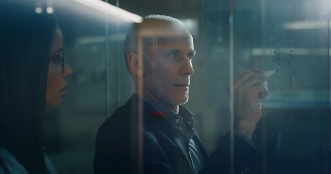 Portrait of CEO manager is explaining to an assistant a strategy of a new marketing for an agency by writing on a glass wall with a marker in an office.
