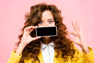 curly girl showing ok sign and smartphone with blank screen, isolated on pink