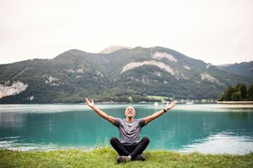 A senior man pensioner sitting by lake in nature, doing yoga exercise.