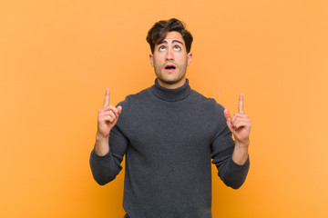 young handsome man looking shocked, amazed and open mouthed, pointing upwards with both hands to...