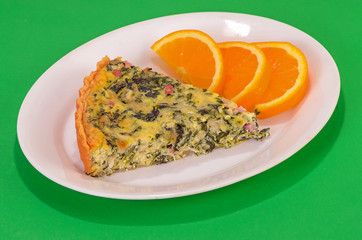 Spinach quiche with ham and cheese on white plate with orange slices against green background.