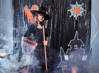 Little serious witch is posing for photographer with broom in the smoke on the Halloween decorations background.