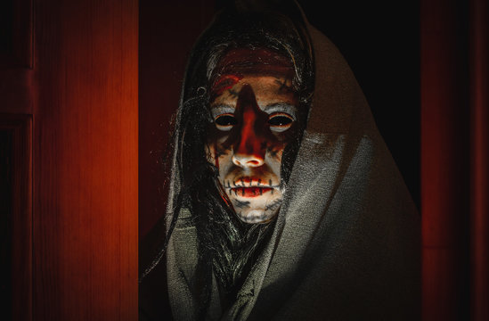 Creepy halloween witch with black eyes, scars on her face in a hood stands near opened doors in the night
