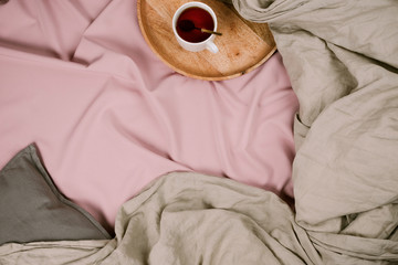 Cozy flatlay of woman's bed with pink sheets and linen grey blanket with tray and tea cup,...