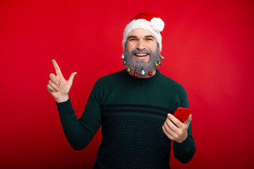 Naklejka premium Cheerful smiling man with decorated beard holding smartphone and pointing at copyspace