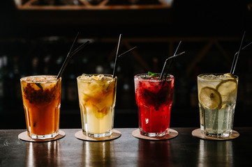 Different types of summer drinks in glasses, cubes of ice and slice of fruits on dark wiooden table.