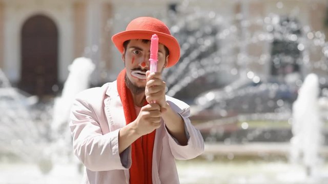 Mime blows soap bubbles on the street at the fountain. Clown shows a sketch in the city.