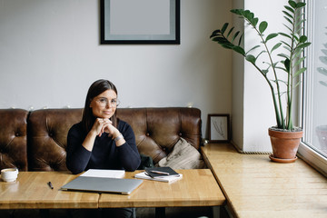 Fototapeta premium Young business woman sitting at table and taking notes in notebook.On table is laptop, smartphone and cup of coffee. Blogger