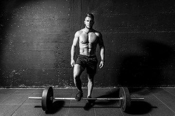 Fototapeta na wymiar Young muscular sweaty fit man with big muscles standing above heavy barbell weight after cross training workout in the gym dark image real people black and white