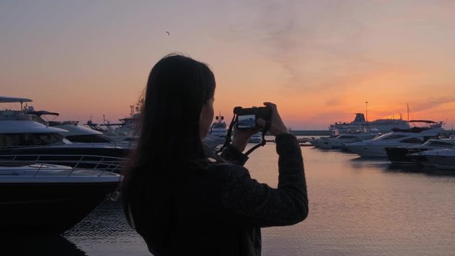 Brunette girl in the seaport taking pictures of yachts on camera at sunset. A woman photographer takes a photo at sunset in the port.