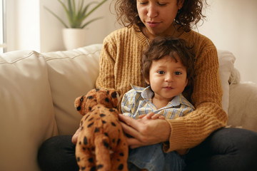 Indoor shot of happy caring young Latin woman sitting on sofa with adorable toddler on her lap playing with stuffed animal spending nice time together. Mother and son bonding in living room