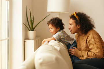 Casually dressed young Latin female with curly hair sitting on couch with her two year son looking...