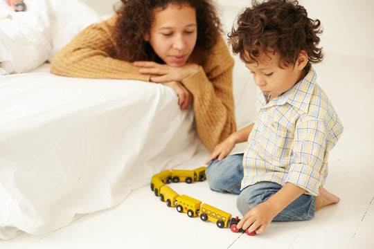Indoor shot of happy young Latin woman in sweater watching her infant son play with toy railway on floor in bedroom, not distracting him. Maternity, child care, early development and imagination