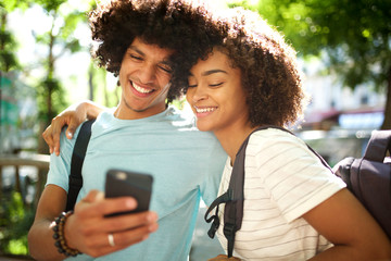 smiling young african american couple standing outside looking at mobile phone