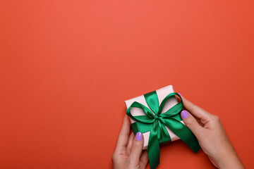 Woman manicured hands holding green wrapped present or gift box on orange brown background with confetti, copy space, top view, flat lay. Background for Valentine's Day, Mother's Day or anniversary