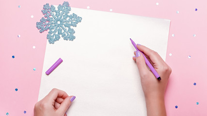 Woman manicured hands holding pen and writing goals and plans for new year. Flat lay on pastel pink desk with confetti and christmas decorations, top view, copy space