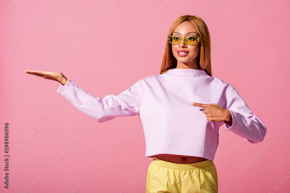 Poster fashionable, smiling african american woman pointing with finger isolated on pink, fashion doll concept