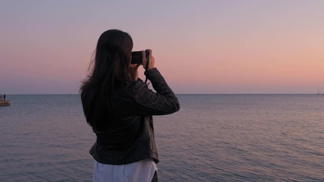Brunette woman at sunset takes a photo on camera standing on the beach. Girl photographer photographing the sea at sunset slow motion.