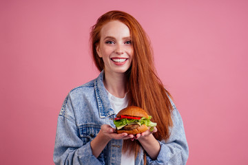 beautiful redhaired ginger redhead woman holding big cheeseburger with cheese,beef cutlet and tomato lettuce in studio pink background